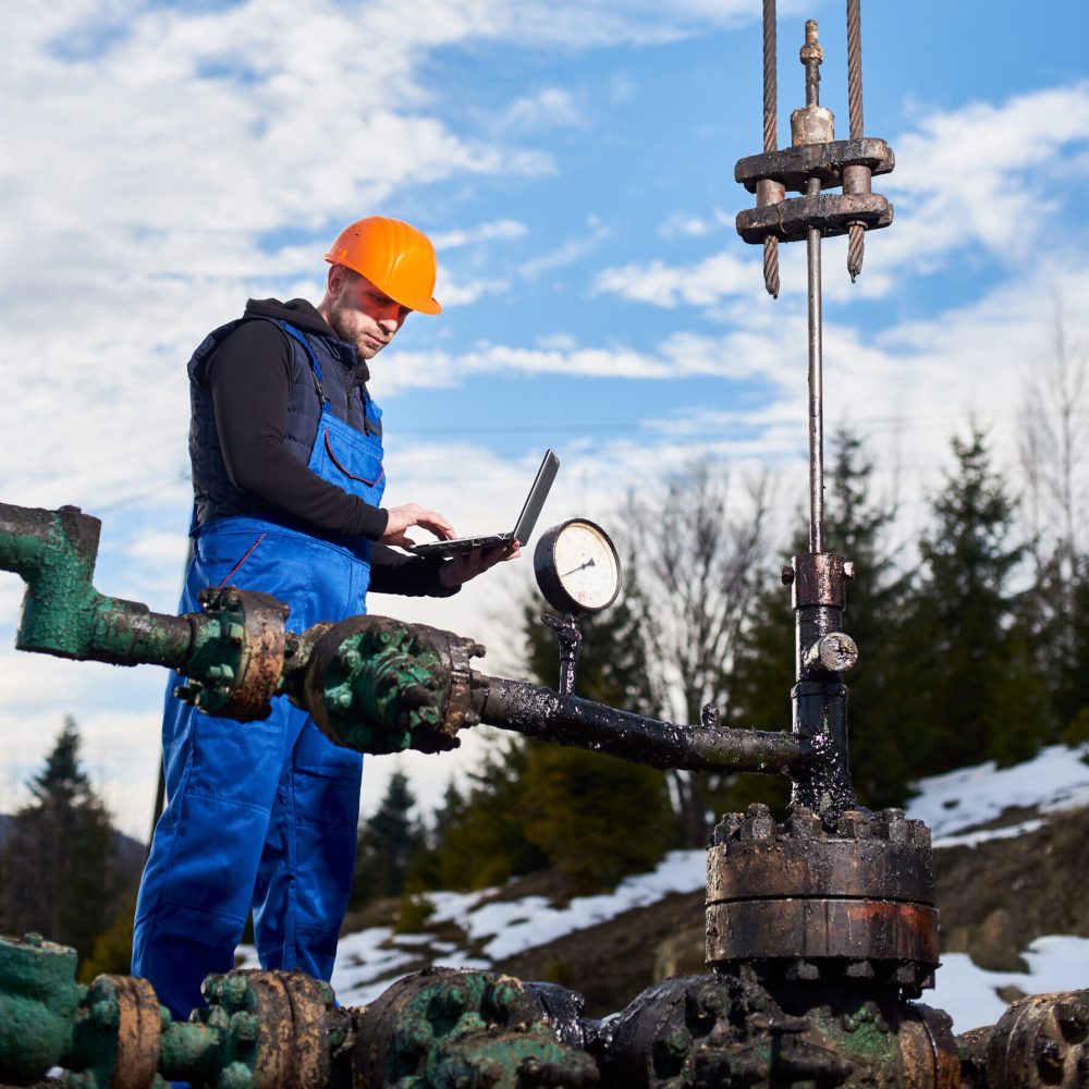 Petroleum engineer using laptop in oil field. Engineer in work overalls and orange helmet checking oil pumping unit at oil field, using laptop. Male worker standing in front of oil well pump jack. Concept of petroleum industry, oil extraction.